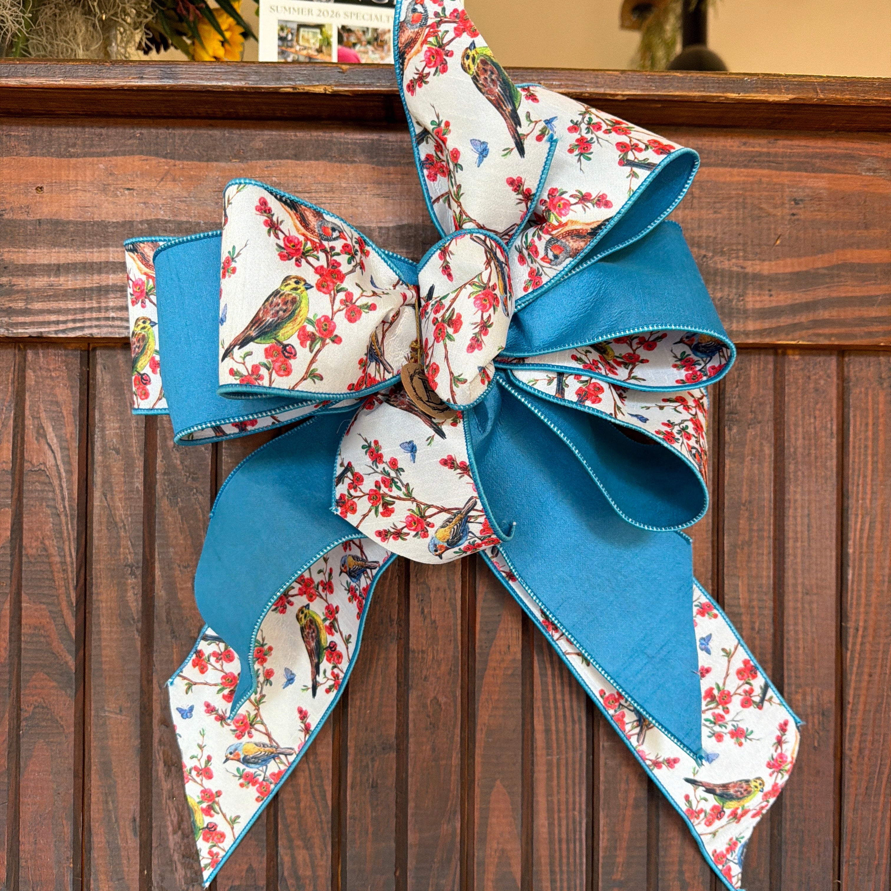 A large decorative bow made from blue satin ribbon and white fabric with a red floral and bird print, tied against a dark brown wooden slat background.