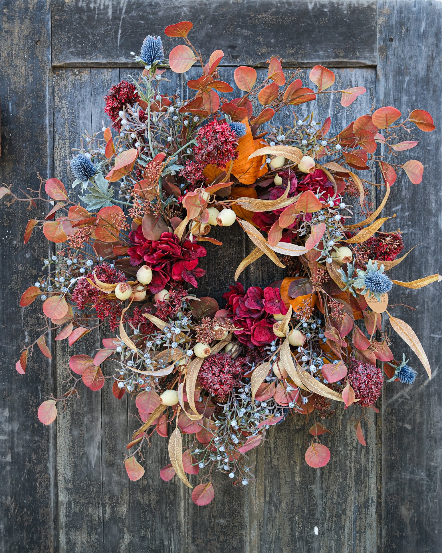 An autumn-themed floral wreath with red and burgundy flowers, orange and rust leaves, blue thistle accents, and beige ribbon elements, hanging on a dark weathered wooden door.