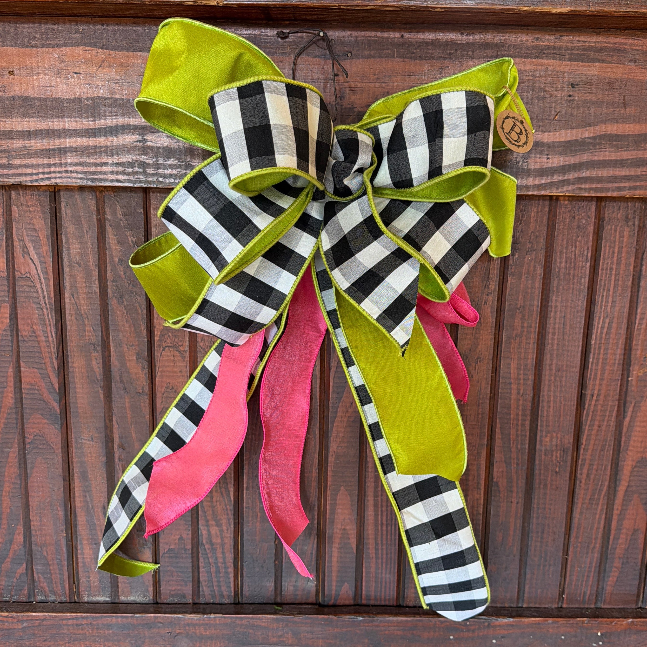 A decorative ribbon bow with black and white buffalo plaid, bright lime green, and hot pink ribbons, hanging on a dark wood-paneled wall.