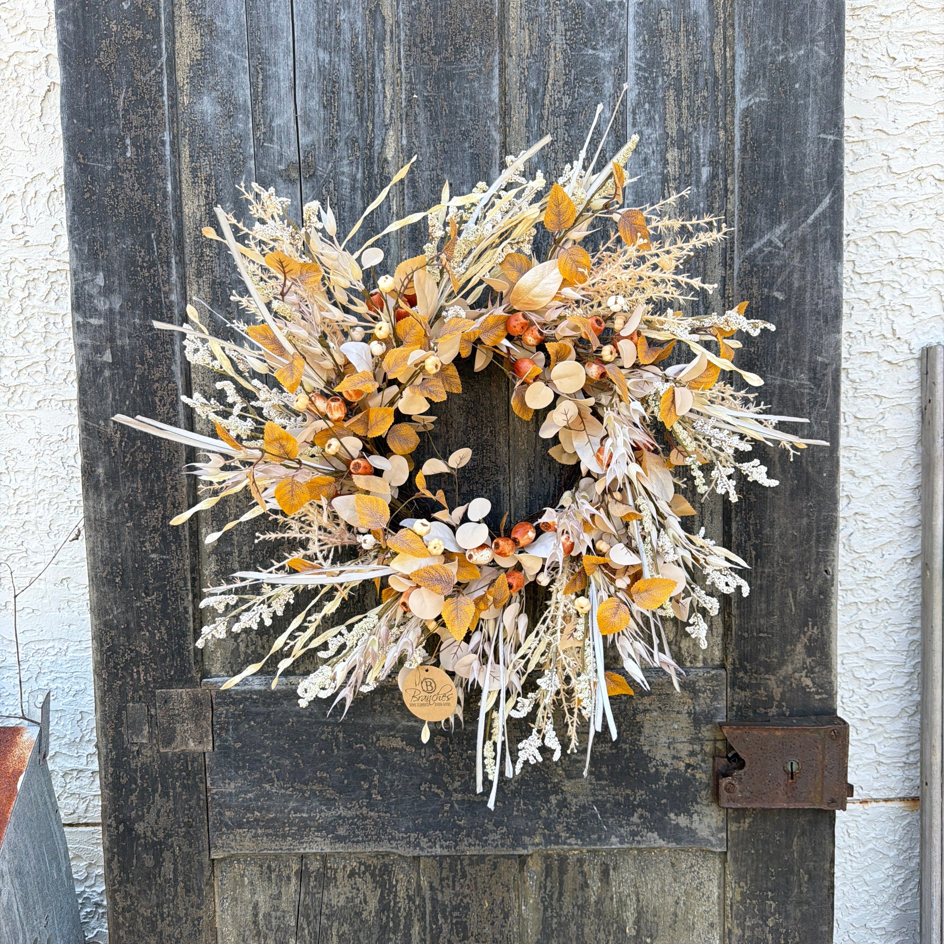 A rustic autumn-themed decorative wreath made of dried beige, brown, and orange foliage, hanging on a weathered dark wood barn door.
