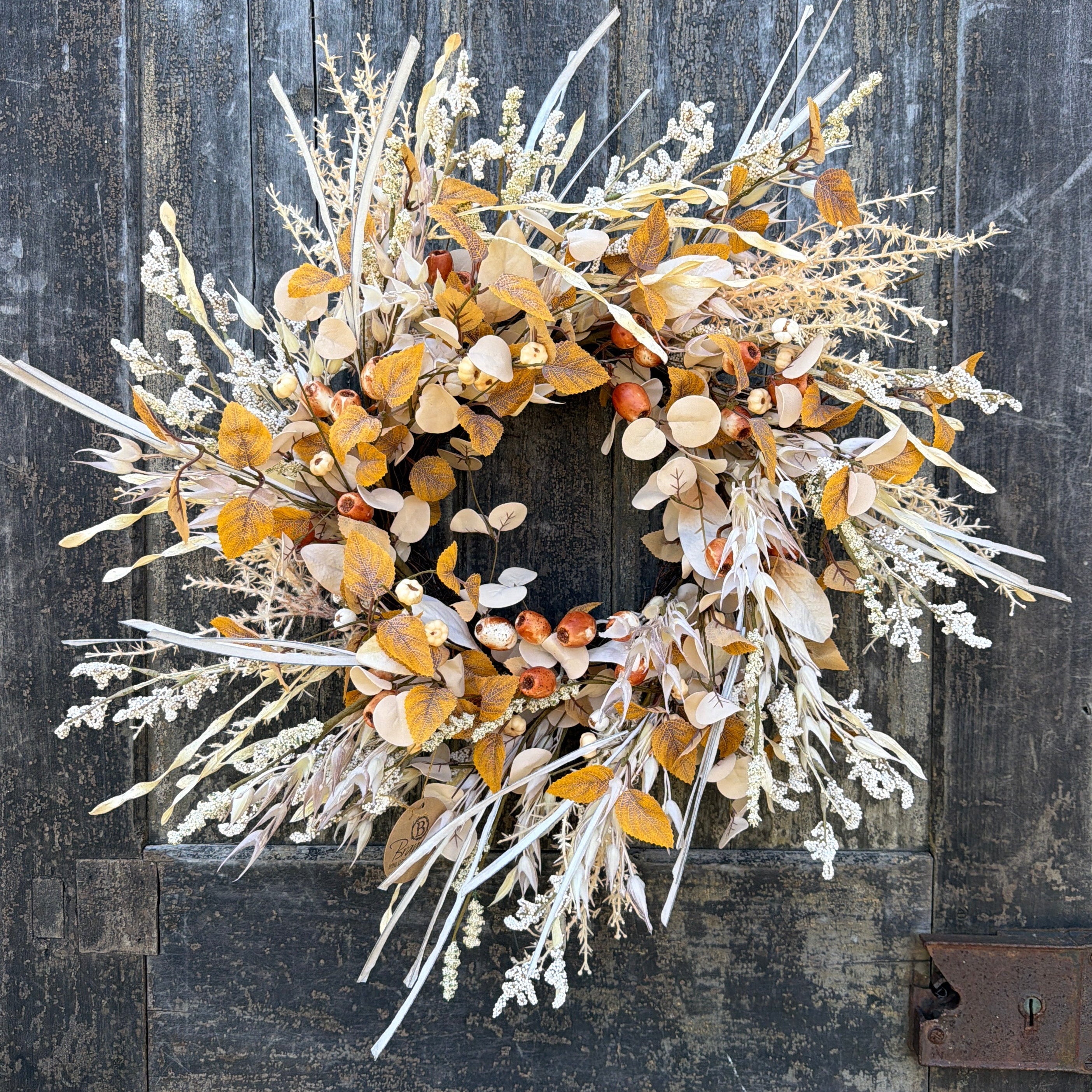 A dried floral wreath with golden-brown leaves, beige pods, and white frosted stems, displayed against a rustic dark gray wooden plank background hilton head island autumn fall
