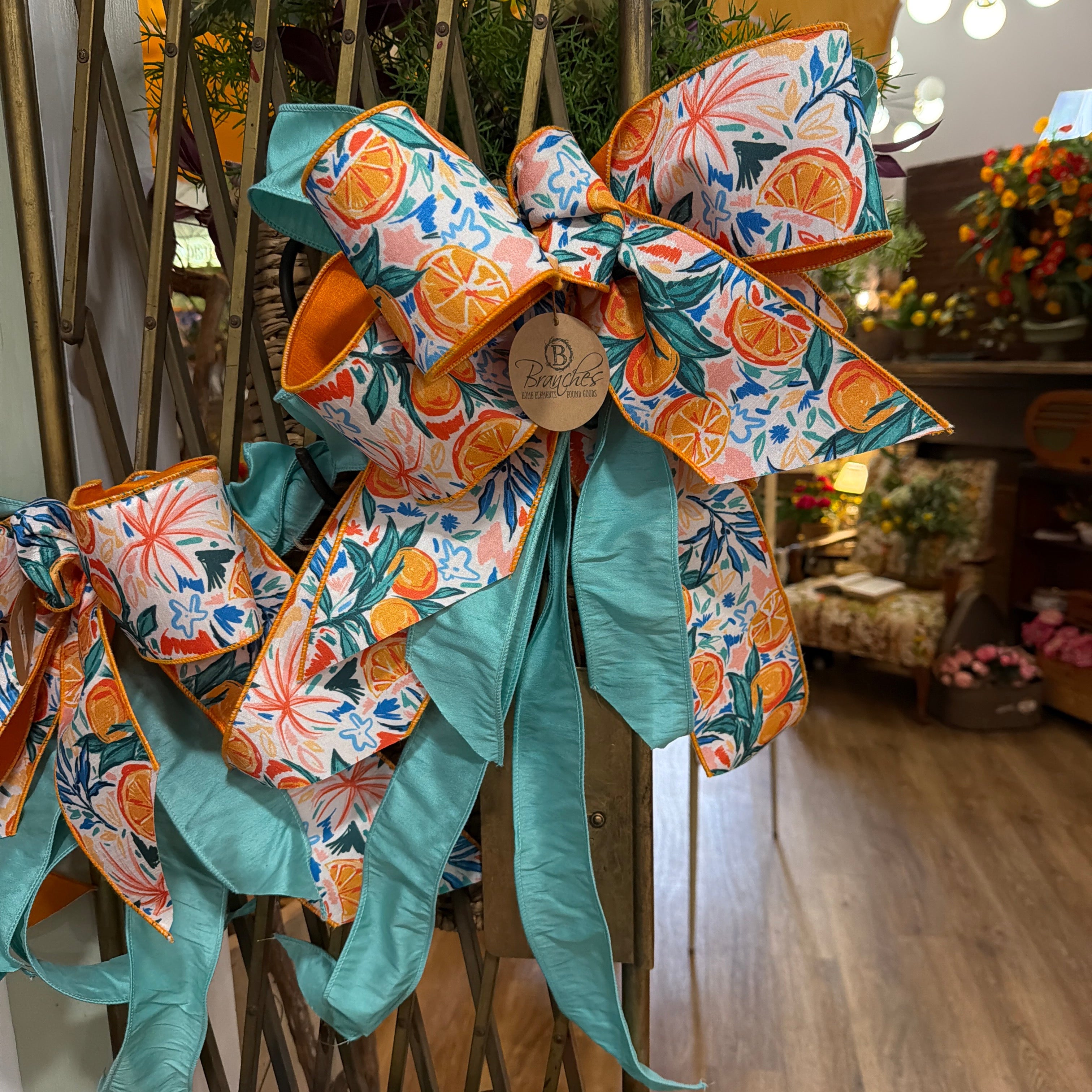 A large decorative bow with a tropical citrus print in orange, teal, blue, and white, accented with a small wooden tag and a green wrapped item on a wooden display.