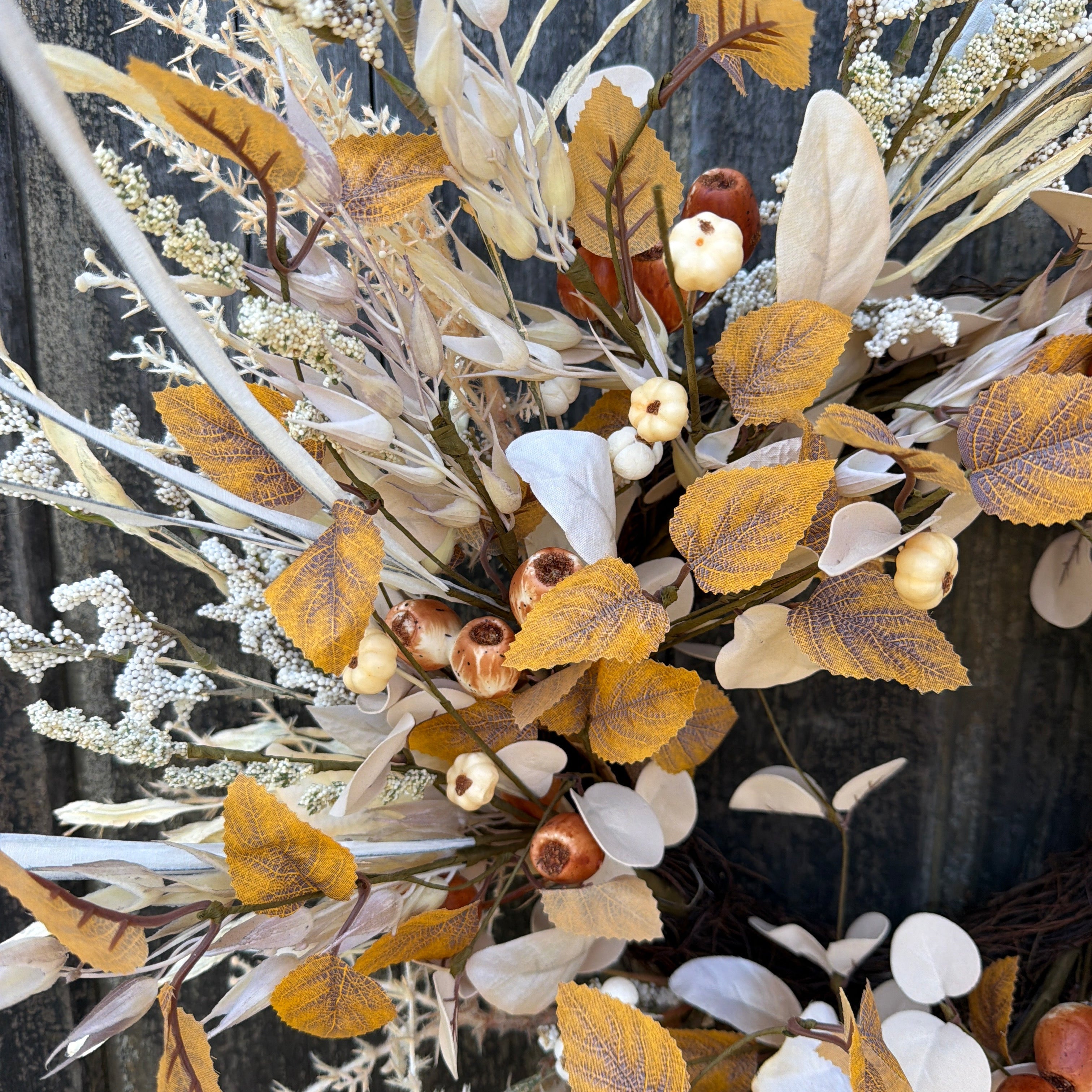 An autumn-themed artificial wreath featuring faux leaves in golden yellow, beige, and brown tones, mixed with white berries, acorns, and dried grasses, displayed on a dark rustic wooden surface.