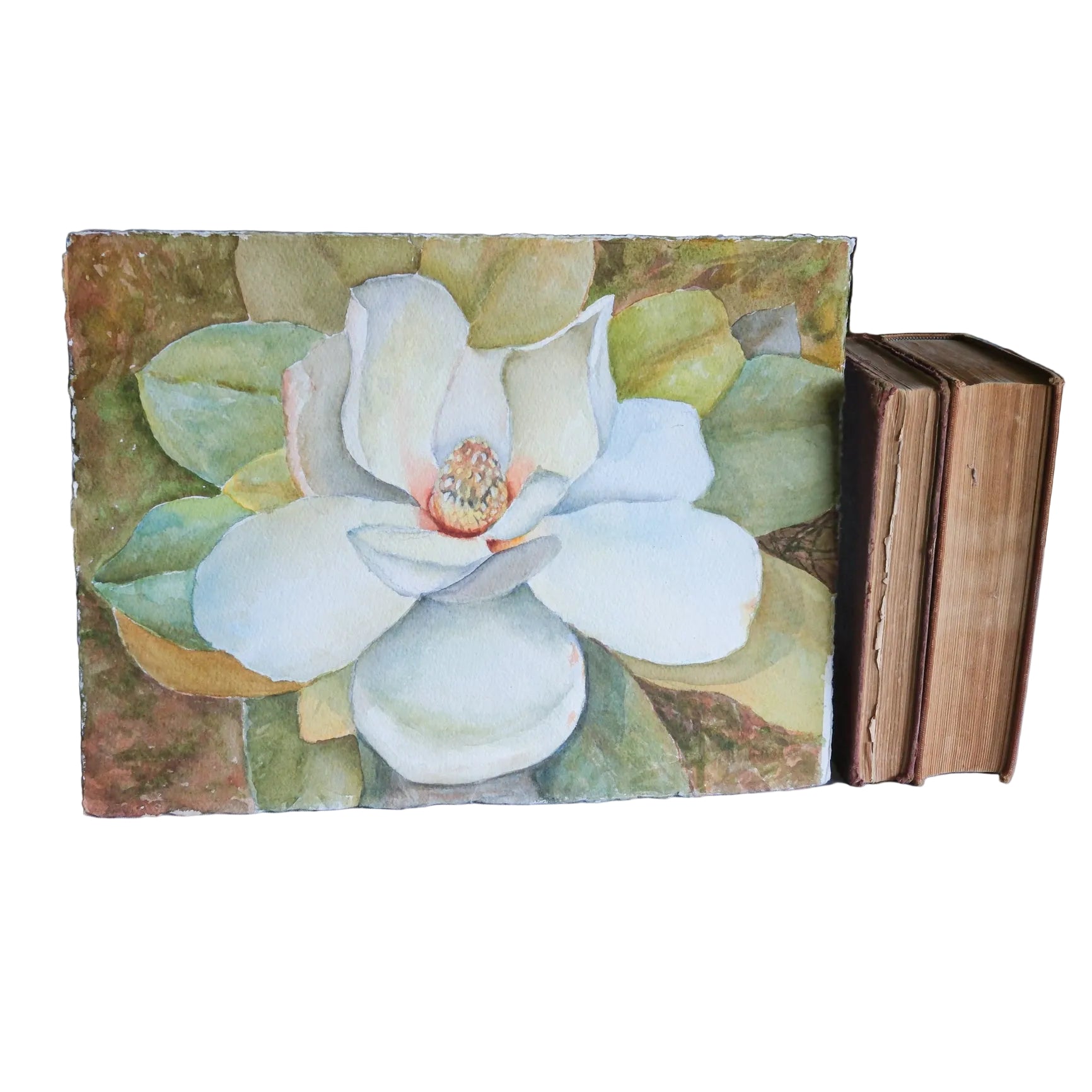A square watercolor painting of a white magnolia flower with green and yellow leaves, displayed on a white fabric-covered surface next to a stack of vintage books.