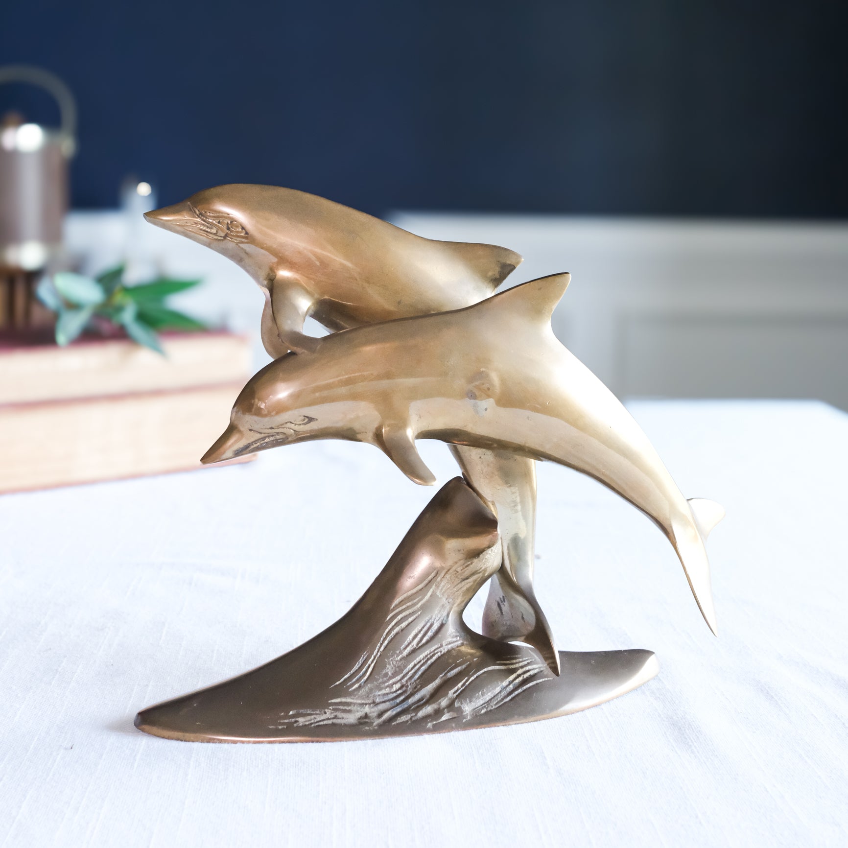 A bronze-finished sculpture of two leaping dolphins rising from a wave-like base, displayed on a white surface with a softly blurred indoor background.